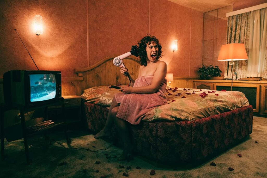 Image of Greta Fernández as Trixie blow drying her hair on a bed in terror