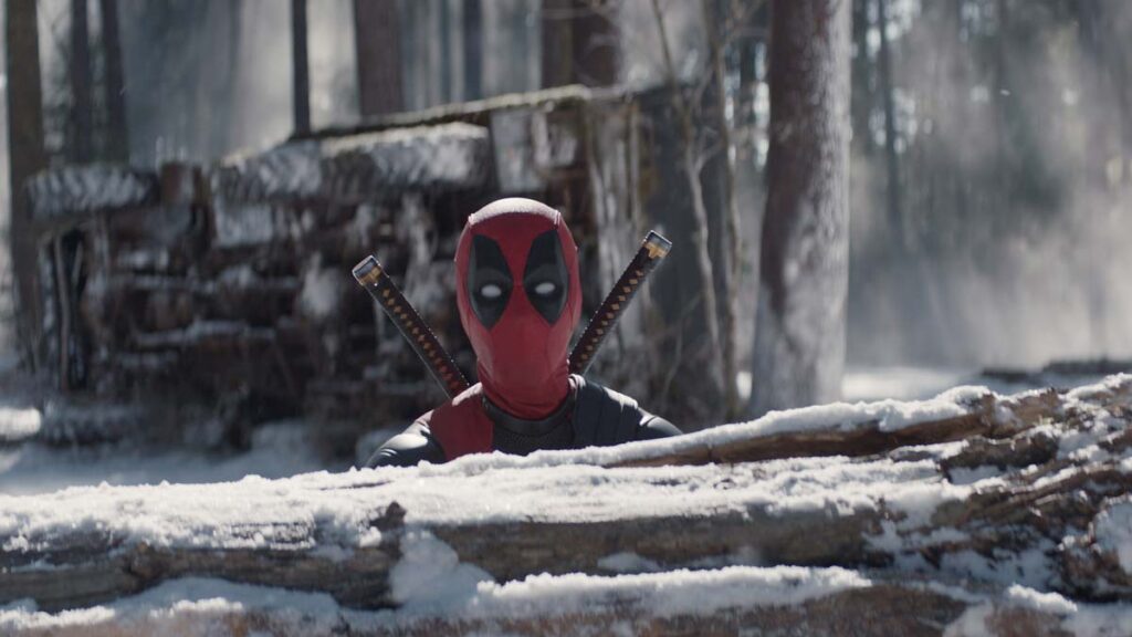 Deadpool pops his head up from behind a snow-covered log.