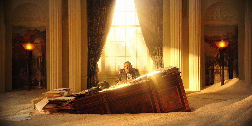 Giangarlo Esposito sits at a desk that is sinking in sand